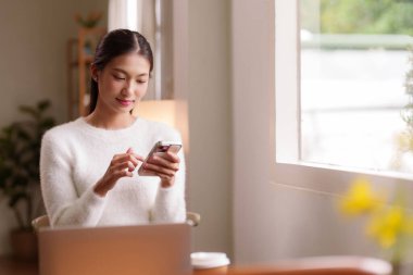 Genç Asyalı bayan ofis masasında parlak pencerenin yanında mobil cihazla etkileşime geçiyor. Laptop görünür. Kapalı bir ortamda rahat bir şekilde bağlanmış gibi görünüyor..