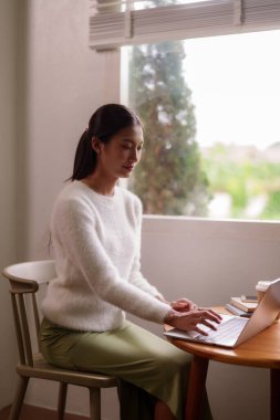 Genç Asyalı kadın evdeki bilgisayarı kullanarak uzaktan çalışmaya odaklanmış. Yumuşak doğal ışıkla yıkanmış rahat bir ortam üretkenliği ve dijital yaşam tarzını geliştiriyor..