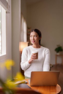 Asyalı kadın ferahlatıcı bir mola veriyor. Sıcak kahve içiyor. dışarı bakıyor. Huzurlu ofisinde ilham buluyor.