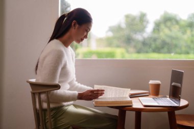 Genç Asyalı kadın kitap okumaya daldı. Kapalı alanda rahat bir ortam hazırladı. Doğal ışık odayı doldurdu.