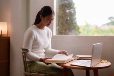 Serene Asyalı kadın evinde parlak bir pencerenin yanında kitap okumakla meşgul. Dizüstü bilgisayarın yanında samimi bir öğrenme deneyimi sergiliyor.
