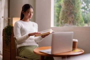 Sakin bir Asyalı kadın rahat evinde kitap okumaktan hoşlanır. Laptop ve kahve rahat bir gün için yakınlarda..