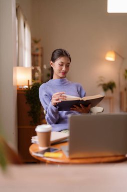 Genç bir Asyalı kadın, rahat evinde dizüstü bilgisayar ve kahveyle çevrili kitap okumaktan keyif alıyor. Huzurlu bir rahatlama ve öğrenim buluyor.