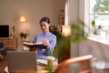 Genç Asyalı kadın evde büyüleyici bir kitap okuyup kişisel gelişim ve rahatlama ortamını teşvik etmekten zevk alıyor.