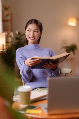 Genç Asyalı kadın rahat bir evde kitap okuyor rahat yetişkin bir kadın evde sessizce vakit geçiriyor ve refahı öğreniyor.