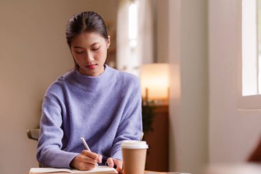 Genç bir Asyalı kadın evde çalışırken defterine yazıyor. Odaklanmış görünüyor ve huzurlu atmosferden hoşlanıyor. Sıcak ortamında bir fincan sıcak kahve var..