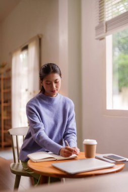 Odaklanmış Asyalı kadın, evinde bir deftere, rahat bir yaşam alanında sakin ve üretken bir sabahın tadını çıkarıyor.