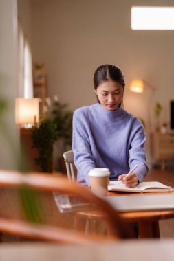 Genç Asyalı kadın, rahat ev ortamından kişisel gelişimi ve yoğun konsantrasyonu teşvik ederek kendi kendine çalışmayla meşgul.
