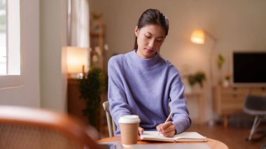 Genç Asyalı kadın evdeki bir deftere yazmaya odaklanmış. Sessiz çalışma, kişisel planlama ya da uzak iş işleri için mükemmel.