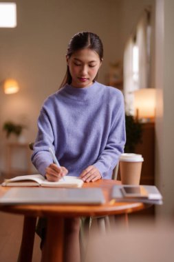 Çalışmaya dalmış genç Asyalı kadın ev ortamında özenle çalışarak kişisel gelişim için notlar alıyor.
