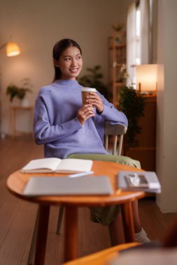 Rahat genç Asyalı kadın evinde ya da yaşam alanında sıcak bir şeyler içiyor. Düşünceli görünüyor. Günlük işine başlayabilir ya da huzurlu bir anın tadını çıkarabilir..