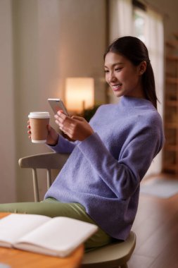 Genç Asyalı kadın evde rahat rahat akıllı telefonlara bakıyor. Rahat bir ortamda kahvesini yudumluyor. Dijital bağlantı eğlencesi