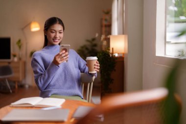 Genç Asyalı kadın, elinde kahve fincanı olan telefona bakarken gülümsüyor.