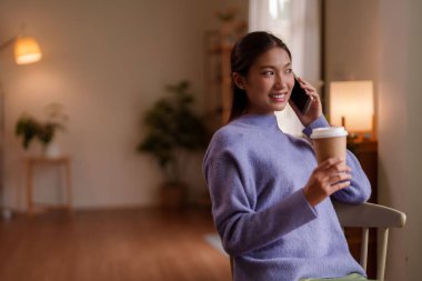 Genç Asyalı kadın telefonunda rahat yaşam alanında sıcak bir kahve fincanı tutarak gündelik bir konuşma yapmaktan hoşlanıyor.