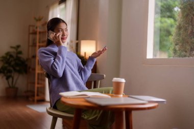 Rahat, Asyalı genç bir bayan ev ofisinden uzaktan çalışırken telefonla konuşuyor.