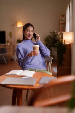 Asyalı bir kadın evde kahve içerken telefonla konuşmaktan hoşlanır. Rahat oturma odası ortamında rahat yetişkin kadın üretken uzaktan kumanda. Dijital iletişim yaşam tarzı