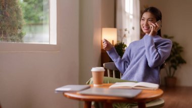 Asyalı bir kadın, parlak bir iç mekanda günlük gezici sohbetten hoşlanır. Sıcak bir içecek ve kişisel aletlerle pencere kenarında dinleniyor.