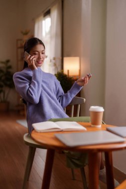 Asyalı dişi kapalı alanda telefonunu kullanarak mutlu bir şekilde iletişim kuruyor. Modern, aydınlık yaşam alanında rahat bir anın tadını çıkarıyor.