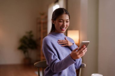 Genç Asyalı kadın elinde bir cep telefonu tutarken keyifle gülümsüyor. Kablosuz bir şekilde kablosuz olarak evinde vakit geçiriyor.