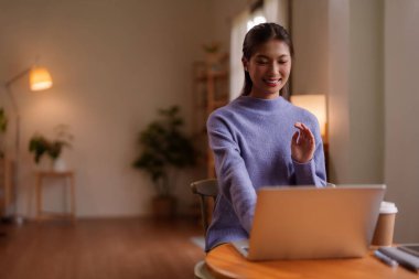 Mutlu Asyalı kadın gülümsüyor. Ev ortamında dizüstü bilgisayarla iletişim kuruyor. Uzak çalışma verimliliği ve dijital bağlantı.