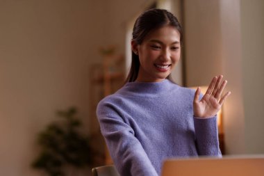 Neşeli Asyalı genç kadın, modern teknolojiyi kucaklayan rahat çalışma alanından uzaktan bağlanan sanal buluşmadan hoşlanıyor.