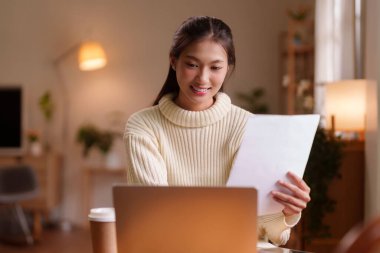 Evde dizüstü bilgisayar ve kağıtlarla çalışan Asyalı kadın, mutlu ifadeyi çevrimiçi öğrenme ortamında verimli uzaktan çalışmalara odaklandı