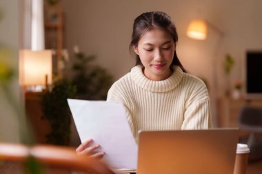 Evde çalışan genç Asyalı kadın. Bilgisayarındaki önemli belgeleri inceliyor. Bu resim odaklanmış ve üretken bir uzak çalışma ortamı yakalar.