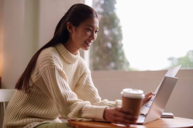 Asyalı genç kadın bilgisayar belgelerine ve evden uzaktan çalışan kahveye odaklanmış rahat bir çalışma alanı yaratıyor.