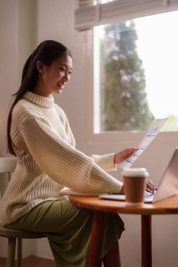 Mutlu Asyalı kadın evdeki bilgisayarla belgeleri inceliyor. İş analizine odaklanıyor. Rahat bir çalışma ortamının tadını çıkarıyor.