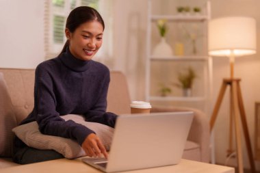 Mutlu Asyalı kadın evde taşınabilir bilgisayarını kullanırken gülümsüyor. Rahat yaşam alanında rahatlık ve verimlilik buluyor..
