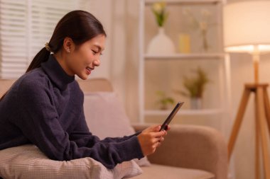 Asyalı bir kadın evde boş vakit geçirmekten hoşlanır. Sosyal medyada internette gezinen mobil bir cihaz tutuyor. Modern yaşam tarzı kavramları için mükemmel..