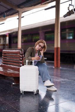 Asyalı yaşlı kadın yolcu tren istasyonu platformunda bekliyor. Akıllı telefon kontrol programı ile bir sonraki bacak yolculuğu seyahati.