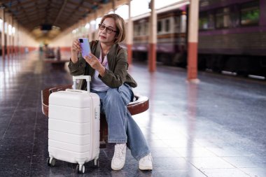 Asya 'nın kıdemli bayan yolcusu tren istasyonu platformunda bekliyor. Mobil iletişim teknolojisini kontrol ediyor. Modern seyahat deneyimi.