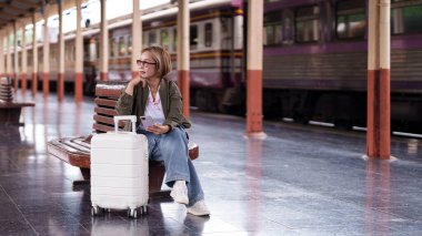 İstasyondaki son sınıf bayan yolcu. Telefonunu kontrol ediyor. Beyaz bavulun yanında, seyahat beklentisi içinde..