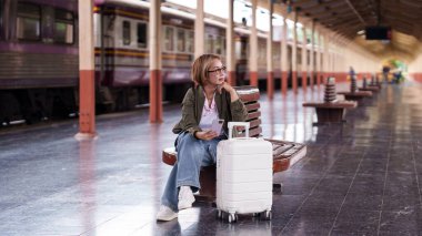 Asyalı yaşlı kadın tren istasyonu platformunu bekliyor. Yolculuğun tadını çıkarın. Telefon, seyahat, macera.