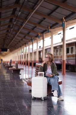 Asyalı yaşlı kadın tren istasyonunda oturuyor. Bavullu telefon bekliyor. Gitmeden önce rahatlatıcı ruh halini yansıtıyor.