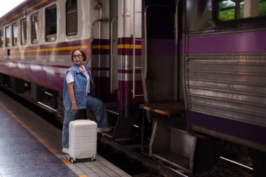 Yaşlı Asyalı kadın, tren istasyonu platformunda beyaz bavul yolculuğunu hazırlıyor. Yalnız başına seyahat ediyor. Emeklilik tatilinin tadını çıkarıyor.