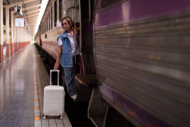 Yaşlı Asyalı kadın bavuluyla tren istasyonunda duruyor. Yolculuğa hazır..