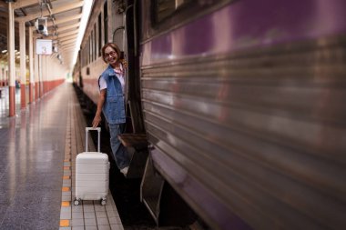Yaşlı Asyalı kadın tren istasyonunda elinde bavulla gülümseyerek tatil için hazır bekliyor.