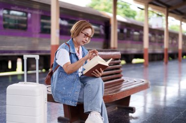 Olgun Asyalı kadın yolcu ahşap bankta oturur tren istasyonunda yolculuk macerası için beklerken roman okur.