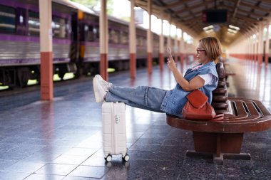 Yaşlı Asyalı kadın rahatlamak için akıllı telefon tren istasyonu platformunu kullanıyor.