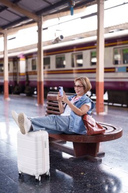 Yetişkin bir Asyalı kadın tren istasyonunda telefonunu kontrol ederken mutlu bir şekilde bavulunu dinlendiriyor ve yolculuğunu bekliyor.