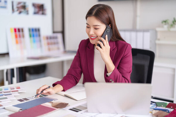 An Asian woman engages a client via phone call reviewing material samples at her design studio. She focuses on creative planning.