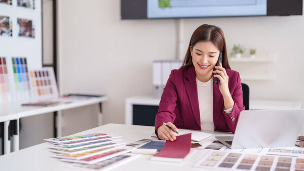 Happy Asian woman interior designer discusses project details on phone. She selects color samples for new home decor in a bright office.