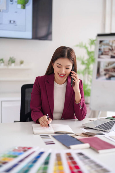 Asian female professional smiles talking phone designing home interiors drafting notes using color swatches in contemporary office setting