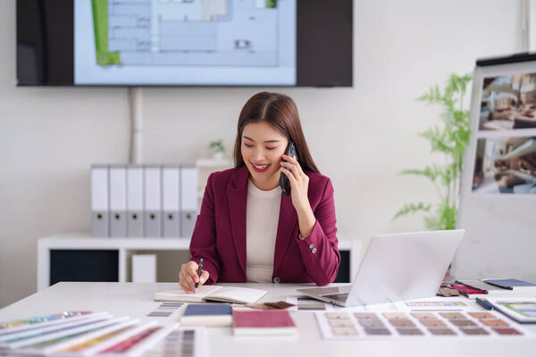 Asian adult professional manages project modern office. Communicates via phone takes notes. Architectural plans material samples display design work.
