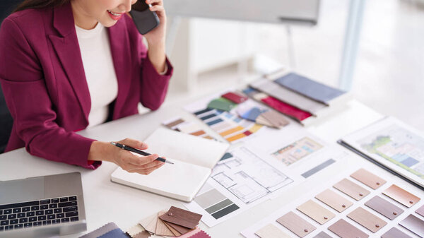 Asian woman interior designer collaborates home projects selects color schemes materials clients uses smartphone laptop manages professional tasks office