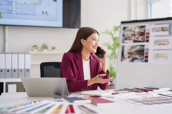 Asian adult woman architect actively engages phone conversation office. Discussing design concepts property solutions client ideas professional workspace.