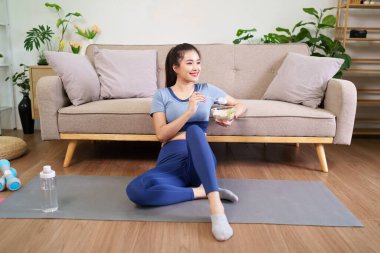 Egzersiz sonrası yoga minderinde besleyici yiyecekler yiyen gülümseyen Asyalı kadın. Etkin sağlığın teşvik edilmesi yerli sağlık yaşam tarzı.