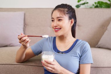 Gülümseyen Asyalı yetişkin kadın evdeki bardaktan besleyici yoğurt yiyor. Sağlıklı yaşam tarzını ve refahı teşvik eder. Rahatlama anı.